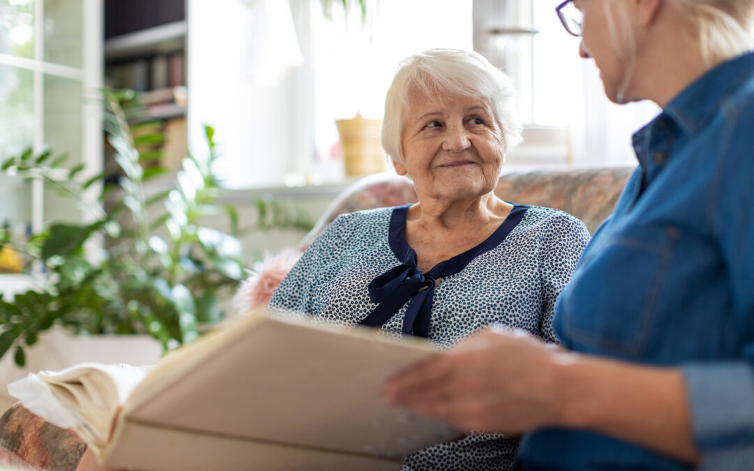 Live-in caregiver seated next to older woman on couch. They are looking at a photo album.