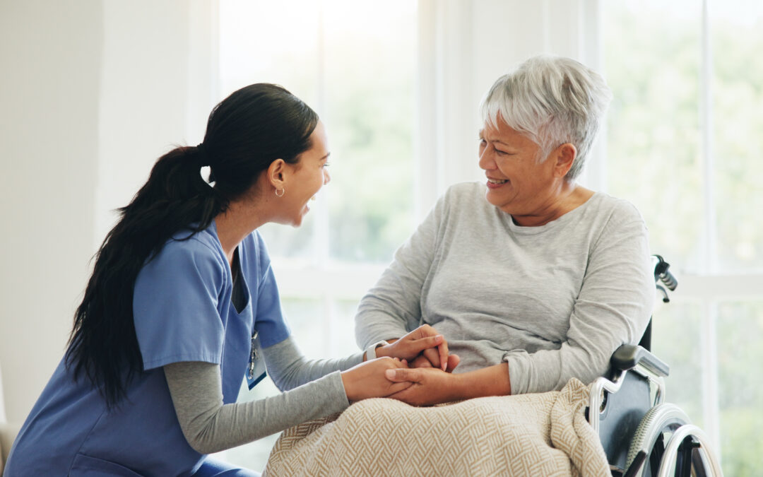 A female caregiver in a blue shirt holds the hands of a senior woman in a wheelchair for heart-of-assisted living blog.