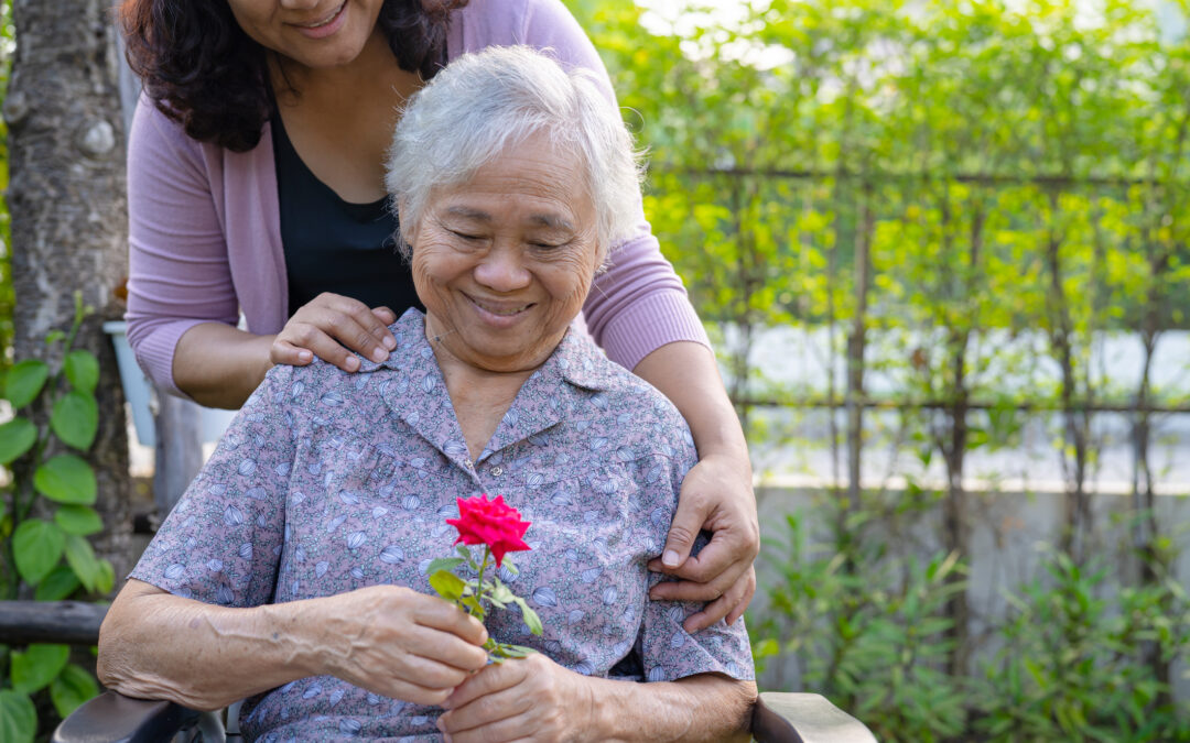 A young caregiver hugs and helps an Asian senior seated in a wheelchair at the park for early-stages-of-alzheimers blog.